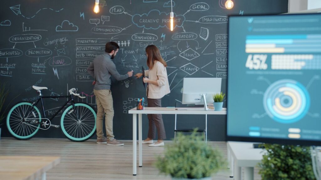 Two colleagues brainstorm ideas on a chalkboard in a modern office setting, discussing strategies and plans.