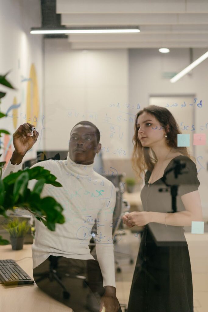 Two coworkers collaborate on a glass board in a modern office setting.