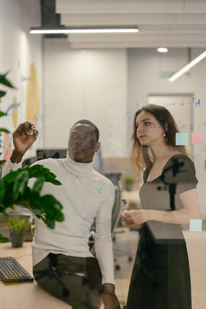 Two coworkers collaborate on a glass board in a modern office setting.
